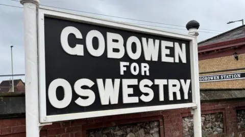There is a sign held up by white pillars and a white frame, but on a black board. On the sign, there are big bold letters, all in capital - saying 'Gobowen for Oswestry.'