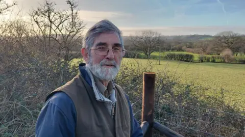 Local Democracy Reporting Service A man with glasses, grey hair and a beard wearing a heavy sweater and a brown gilet stands before green countryside dotted with trees and hedgerows. 