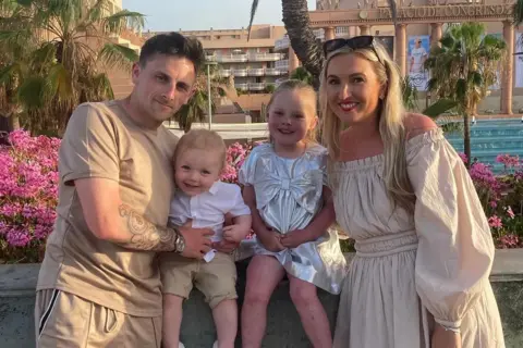 Jodie McCann A man in a tan t-shirt, with a tattoo on his arm, and a woman in a white dress and with sunglasses on her head, stand on either side of two children - both very young - while outside what looks like a hotel.