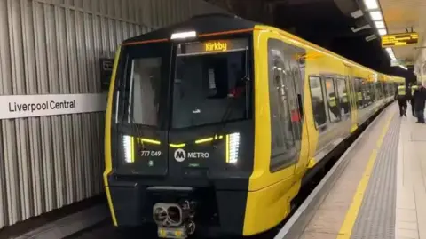 A yellow and black Merseyrail train at a platform at Liverpool Central station. The destination Kirkby is displayed in yellow at the front of the train which carries the Metro logo.