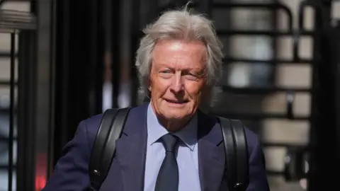 PA Media Paul Tweed. He has grey hair and is wearing a navy suit, blue shirt and navy tie. He has a black backpack on and the straps on his shoulders are visible.