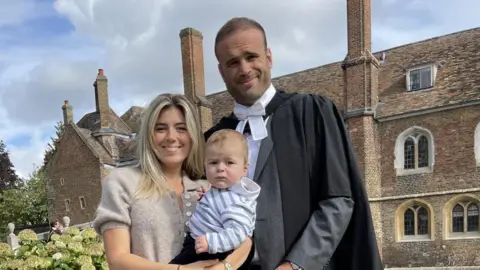 Jamie Roberts A couple, Jamie Roberts and his wife Nicole, stand with their baby in front of Cambridge University.
