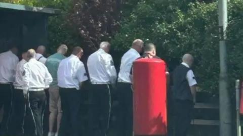 Social Media A number of men are standing near a fence with their backs turned appearing to be urinating.  They are standing next to a red post box and what appears to be a bus shelter
