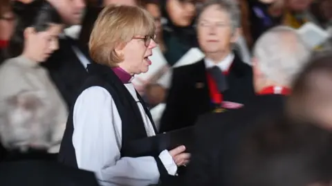 PA Media A woman, Dame Sarah Mullally, stood in the middle of a crowd in a church. She is reading and singing from a hymn sheet.