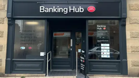 Exterior of a banking hub shop front. The paintwork is black with glass windows. The sign Banking Hub is at the top of the shop front alongside a sign saying Post Office.