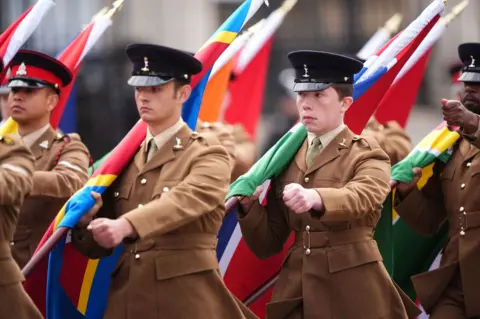 PA Media Members of the procession carry flags of the Commonwealth nations while wearing brown uniforms and black hats, with their fists raised as they march in unison.