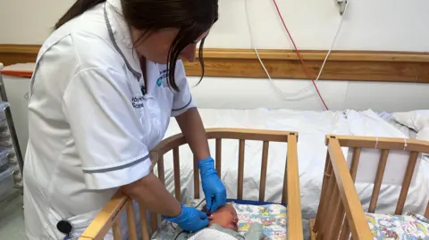 a woman in a nurse's uniform and gloves reaching into a cot 