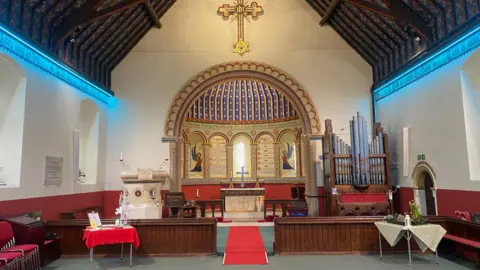 Vernon Lidstone The altar and font section at the front of a church, where there is an organ. There is a cross above the dome-shaped chancel, which is very colourful. The walls are white and red and there is a red rug in front of the alter and a red tablecloth on the table to the left of the image. 