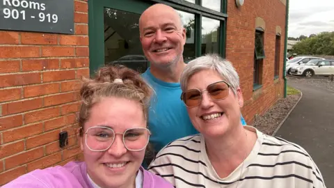 (L-R) Gen, Stephen and Alyson Portlock stood outside a brick office building. Gen wears large square glasses and a pink jumper, Stephen wears a blue t-shirt and Alyson wears a black and white striped top. 