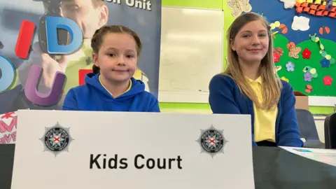 BBC Eva (left) and Layla (right) are sitting beside each other at a table in a classroom. There is a sign on the table with PSNI symbols which says 'Kids Court'. Their uniforms are royal blue and yellow.