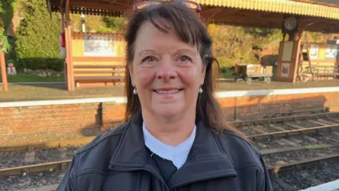 BBC A woman with a dark top is smiling at the camera. Rail track and the opposite platform featuring benches and a clock are visible.