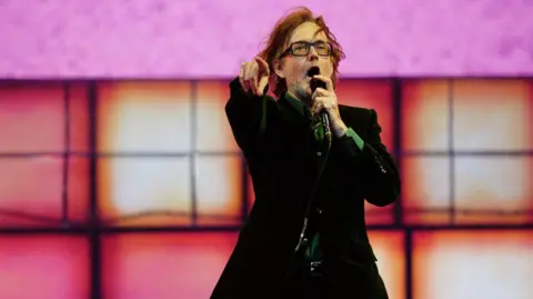 Jarvis Cocker, wearing glasses and a brown jacket, stands on stage pointing his finger and holding a microphone.