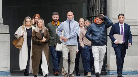 PA Media A group of nine people, including four women and five men, cross a stone square with sombre, determined expressions. 