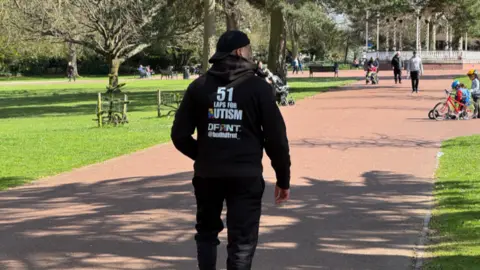 A man in a black hoody with the words 51 laps for autism DFRNT @builtdfrnt is walking away from the camera and appears to be in a park