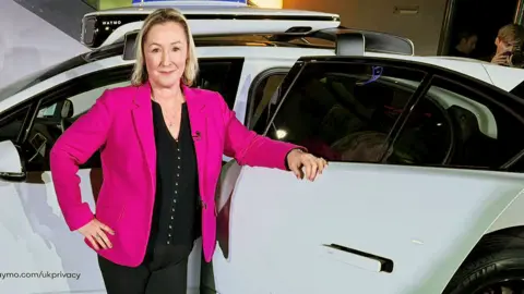 BBC technology editor Zoe Kleinman stands in front of a white car, wearing a red jacket over a black top and black trousers