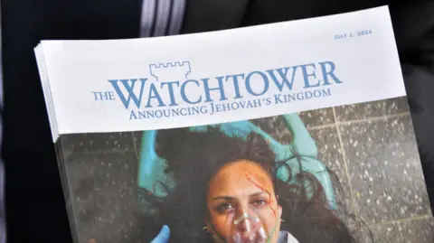A Jehovah's Witness disciple holds out copy of The Watchtower publication (for passers-by) outside Oxford Circus underground station, 8th July 2014. The magazine has a white header but the page is dominated by a woman lying on a stretcher. 