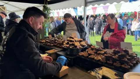 A man with dark hair in a dark coat standing behind a stall loaded with cakes and a man and a woman wearing coats looking at them with marquees and more people in the distance
