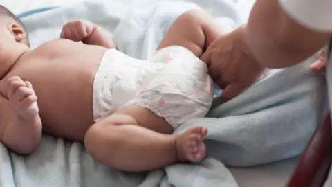 A baby is lying on a towel while having a nappy changed by an adult.
