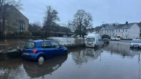 Richard Brassett Vehicles in Lostwithiel parked in flooded water. There are cars and vans parked along the side.