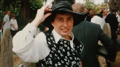 An older family photo of Patricia Grace, it looks like she's at an event, potentially a wedding - there a people in the background in what looks like a churchyard. Patricia wears a black wide brimmed straw hat, she has one hand up to the hat holding the brim and has paired it with a white long sleeved collared blouse and a black smart vest with a white floral print on it. 