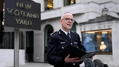 Reuters Sir Mark Rowley, a man with short grey hair and black glasses, stands in front of New Scotland Yard in his police uniform holding a black folder and speaking into microphones.