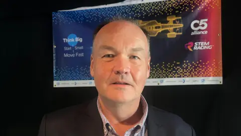 A headshot of a man wearing a checked white and grey shirt with the top button undone and a black blazer. He is looking into the camera with a black wall behind him and a purple and red banner with a graphic of a car behind him, saying STEM RACING.