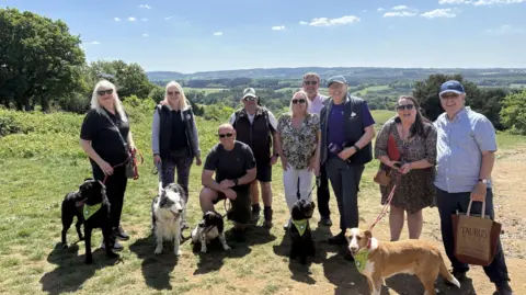 A group of nine people stood in the countryside on a sunny day, with a green vista behind them. One man is carrying a microphone and there are five dogs on leads.