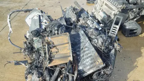 Damage of electrical items caused by fire - a cluster of grey coloured items burnt up in a pile. They have been placed on the ground.