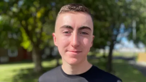 Ryan Dolan, 15, smiles gently at the camera, standing outside with trees in the background. The teenager has dark brown hair and is wearing a black sports t-shirt.