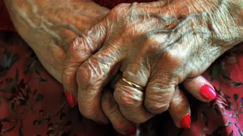 An elderly person's hands are clasped together. Veins are prominent on the hands and the skin has creases on it. The finger nails are painted pink and there is a gold ring on one finger.