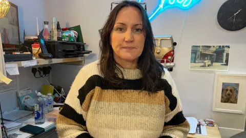 Rhian Synnot is waring a jumper inside her Cain shop, which has various items hung on the wall. Behind her is a desk with office equipment