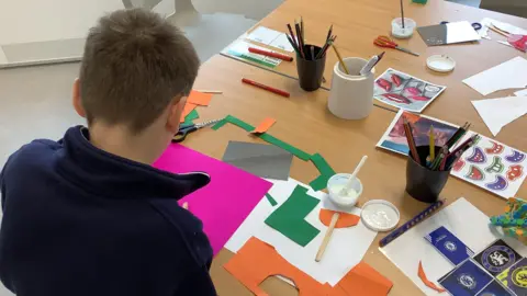 A child sitting with it's back to the camera so you can not see their face. They are sitting in a classroom at a school desk cutting out paper shapes on coloured cards. 