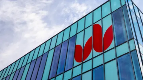 The red University of Bedfordshire logo, which resembles tulips, is printed on the side of a blue glass fronted building