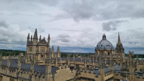 WEDNESDAY - The roofs of Oxford are photographed from roof height against a grey sky. The buildings are made from a yellow stone with grey roofs. Each building has several stone spires. In the distance, the domed roof of the Radcliffe Camera looks striking. On the horizon you can see green trees and overhead the sky is full of light grey clouds.