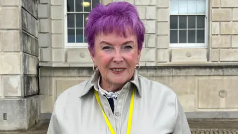 A woman with purple hair and tan coat smiling at the camera, she has a yellow lanyard