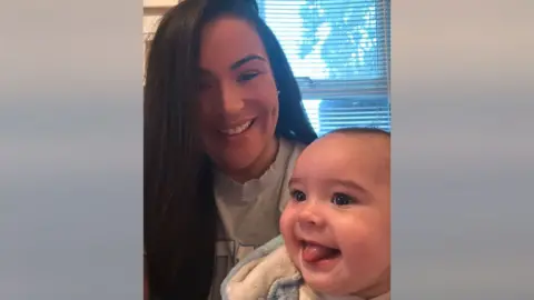 Sasha Field with long black hair, who is smiling as she is holding a baby who is also smiling with his tongue sticking out.
