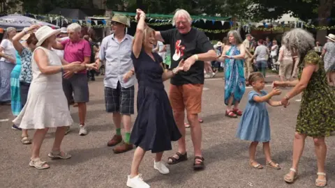 A number of people dancing in the street. Some people are wearing dresses and others are wearing hats.
