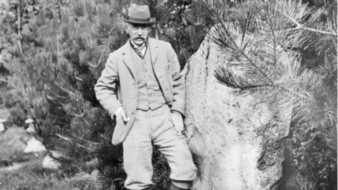 Dorset Museum & Art Gallery / Dorset History Centre Black and white photo of Thomas Hardy posing next to one of the large, wide sarsen stones which is nearly as tall as him. He has a large moustache and is wearing a trilby hat, a tie, waistcoat and jacket and his hands are in his pockets.