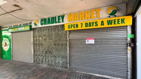 BBC The frontage of the Cradley Market store, with green and yellow signage, with silver shutters that have been closed