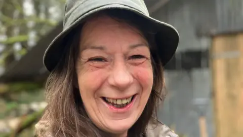 Woman smiling at the camera wearing a waterproof hat in front of the barn where she found the painting