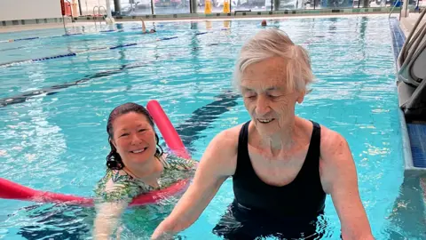 A woman in a black swimming costume lowering herself into a swimming pool which is streching out behind her. She has short white hair and there is a carer in the water, to her left.
