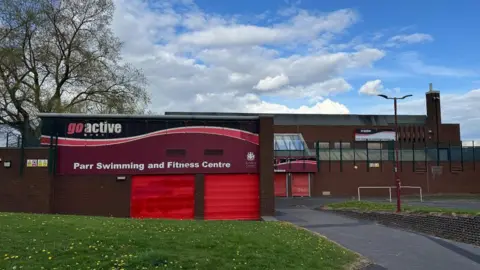 Two brick adjoining buildings with red shutter entrances on then. A sign reads 'Parr Swimming and Fitness Centre'. A patch of grass can be seen in front of it. 