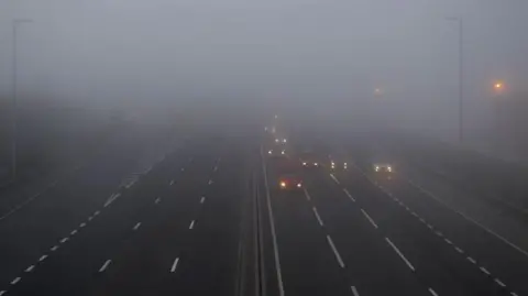 Getty Images Cars driving through fog on a motorway. There are three lanes of traffic on either side and the headlights of the vehicles are fuzzy and some are barely visible due to the foggy conditions. 