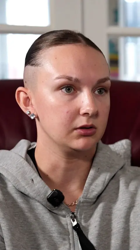 A young woman sits indoors, shown from the shoulders up. She has light skin and green eyes, with her brown hair shaved close on the sides and slicked back on top. She wears a grey zip‑up hoodie and small square stud earrings. A black microphone is clipped near her collar. The background is softly blurred, showing a maroon chair and bright daylight through a window.