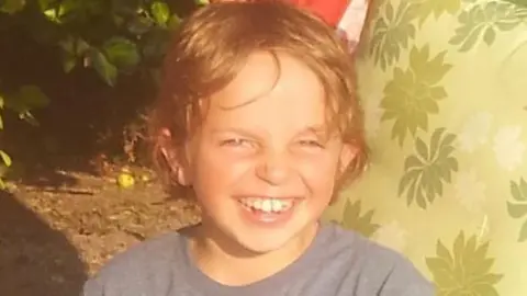 Finlay Church, a young boy is in a dark blue tshirt is smiling at the camera. Behind him there is greenery and on the right of the photo is a green cushion with a leaf pattern on