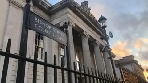 The outside of Bishop Street Court House in Londonderry. A street lamp and black gate is just outside a pillared building.