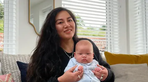 Serena is sitting on a sofa in a bright living room holding baby dressed in light clothing, with large windows, cushions, and a coffee table in the foreground.