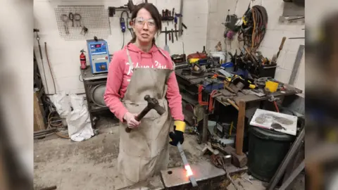 Sally Millington A woman in a pink hoodie and grey apron wearing gloves and googles hammers hot metal in a workshop