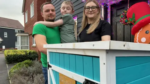 BBC Tom, Evan and Laura Winter stand behind a blue honesty box in front if their home which is decorated for Christmas.