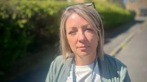 Detective Helena Scott looks into the camera, standing on the kerb with a path stretched out behind her, and a hedge. She wears a white t-shirt, a pale blue/green bomber jacket and a blonde bob with a pair of glasses on her head.
